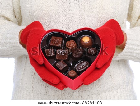 Woman wearing red gloves and white sweater holding a Box Heart of Valentines Day Candy in front of her torso. Close shot in horizontal format.