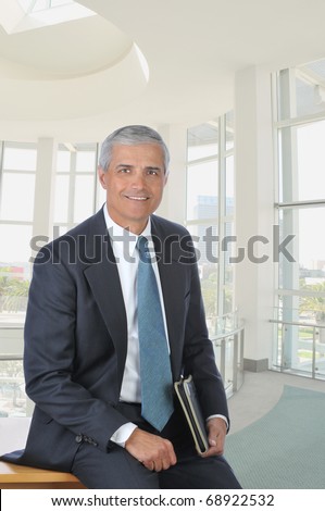 Middle aged businessman sitting on the corner of his desk holding a folder. Vertical format in a modern office building.