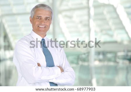 Smiling middle aged businessman standing in the lobby of a modern office building. Man is wearing white shirt and necktie with his arms crossed. Horizontal Format.