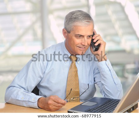 Middle aged businessman with laptop talking on telephone in modern office setting. Square format.