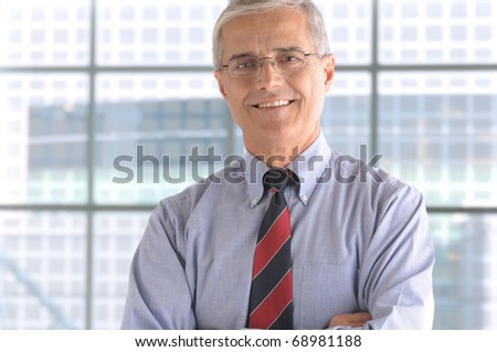 Close up of a smiling middle aged businessman in the lobby of a modern office building. Man is standing in front of a large window wearing a blue shirt and necktie. Horizontal Format.