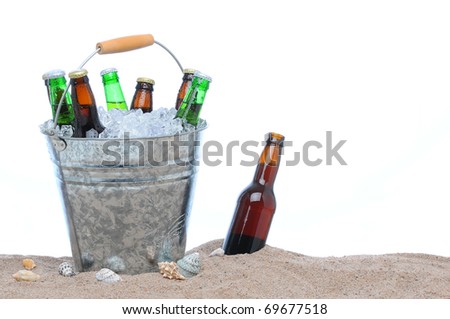 Assorted beer bottles in a bucket of ice in the sand isolated on white. One beer bottle without a cap is by itself stuck in the sand next to the pail.
