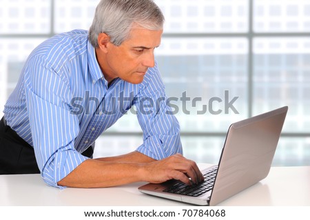 Smiling Middle Aged Businessman leaning on desk using laptop computer with large window background