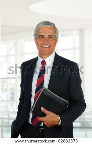 Smiling middle aged businessman in modern office setting holding a small binder. Vertical Format.