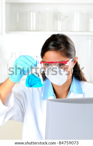 Young female Lab Tech seated behind computer monitor holding up a flask of blue chemicals in laboratory setting. Vertical format.