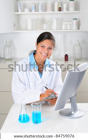 Young female Lab Tech seated behind computer monitor writing notes on a clip board in laboratory setting. Vertical format.