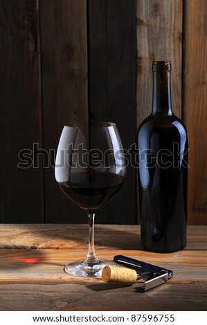 A single bottle of red wine and a wineglass in a rustic wine cellar setting. Directional light with wine glass reflection in table top.
