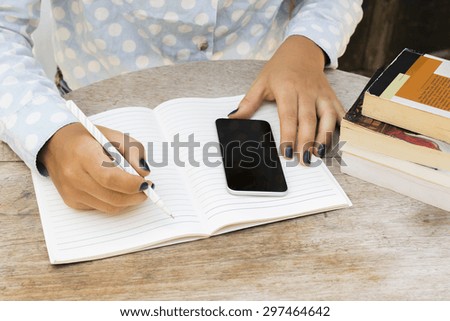 girl writes in a notebook, with cell phone and books on the table