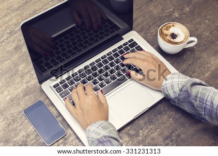 Girl typing on a laptop on a wooden table, with cell phone and cup of coffee