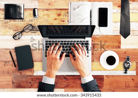 Typing businessman with laptop, cup of coffee, blank diary cover and glasses on wooden table, mock up