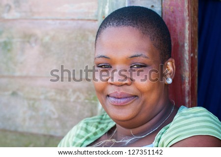 A young African woman, back against the wall and smiling at the photographer.