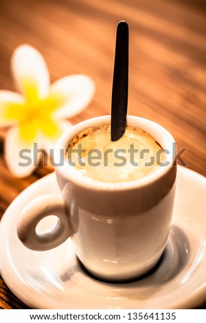 a close-up of a single macchiato stirred on a wooden table with a frangipani in the backround