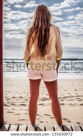 a beautiful young caucasian woman standing  with her back facing front on beach a  cabin deck looking out on a sunny beach day
