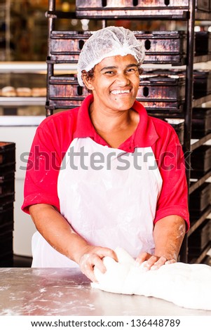 african colored baker smiling proudly while kneading dough on a work counter in a hairnet and plastic apron