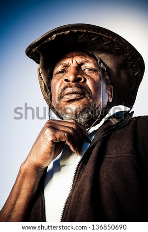 old african farmer in a dark hat frowing down in thought playing with his grey beard
