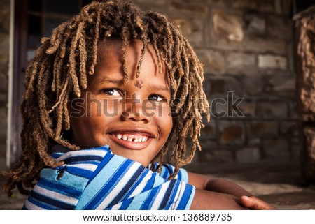 young african boy with rasta hair in a blue striped shirt sitting on a stone porch smiling up in admiration