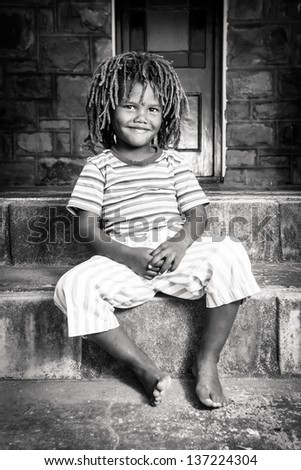 black and white image of a cute young african boy with dreadlocks sitting on a stone porch