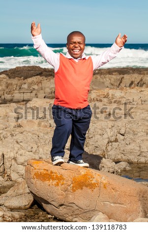 dwarfish african man throwing his hands up laughing openly with a rocky beach in the background