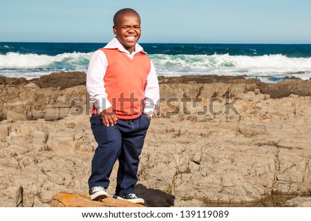 Dwarfish african man standing on a big boulder on the rocky beach in stylish clothes smiling whole heartedly