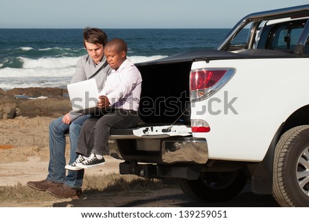 young caucasian man and dwarfish african man sitting on the back of a pickup truck working on a laptop