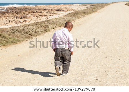 dwarfish african man walking down a dirt road along the coast with his head bent down