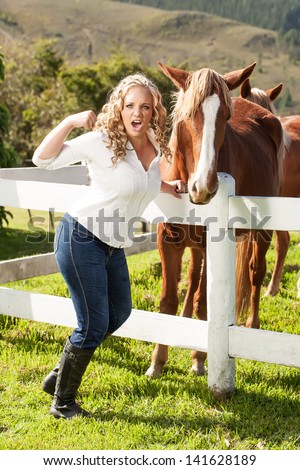an angry teenage caucasian girl with blond bouncy hair getting ready to hit a mahogany colored horse on the other side of the white picket fence