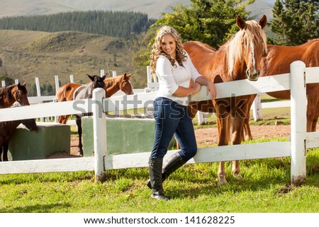 a beautiful caucasian girl with blond curly hair standing against the white picket fence with a mahogany colored horse equally interested