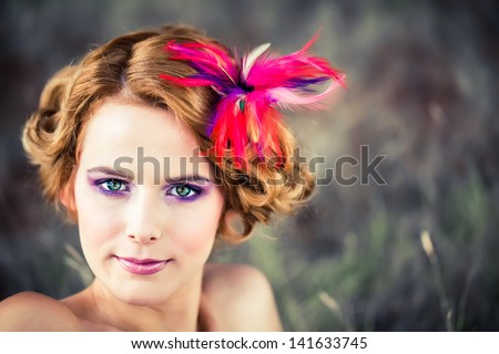 beautiful caucasian model with red curly hair pinned up with a feathered flower sitting in the tall grass