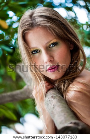 portrait of a beautiful fair haired young model with slight freckles in her face holding on to a tree branch and looking back intensely