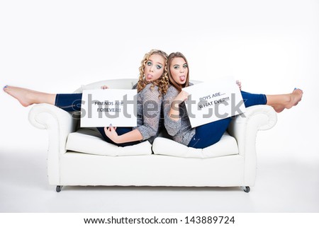 two caucasian friends sitting with their bare feet over a big white couch sticking their tongues out in attitude wearing matching gray tops