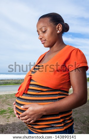 portrait of a young pregnant girl wearing a bright orange striped shirt holding her big belly lovingly and smiling slightly with the ocean in the background