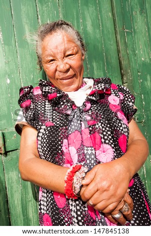 portrait of an old african beggar leaning against a green wooden door with her hands folder in front of her and smiling cutely