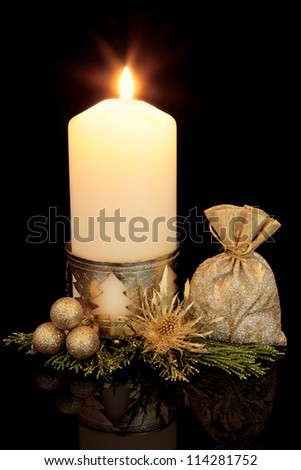 Christmas decoration of gold bauble cluster, thistle flower head, gift bag, leaf sprigs and lit candle with holder over black background.