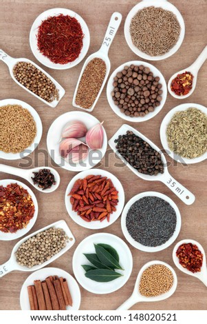 Large spice and herb selection in white china bowls and measuring spoons with millilitre measurement over papyrus background.