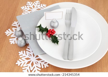 Christmas dinner place setting with white plates, cutlery, name tag, holly, silver bauble and snowflake decorations over oak table background.