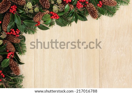 Christmas background  border with holly, ivy, fir leaf sprigs, pine cones over light oak background.