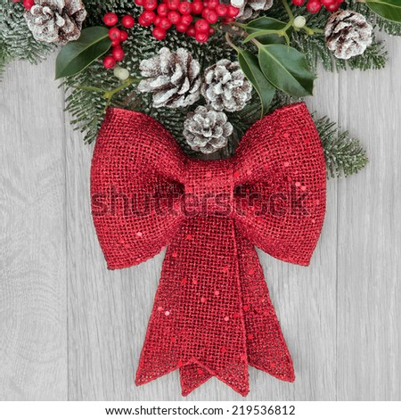 Christmas background  border with red bow decoration, holly, mistletoe, winter greenery and snow tipped pine cones over light oak background.