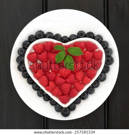 Raspberry and blueberry fruit in a heart shaped bowl on a white round dish over black background.