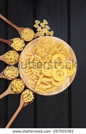 Pasta selection on a wooden board and in olive wood spoons over dark wood background.