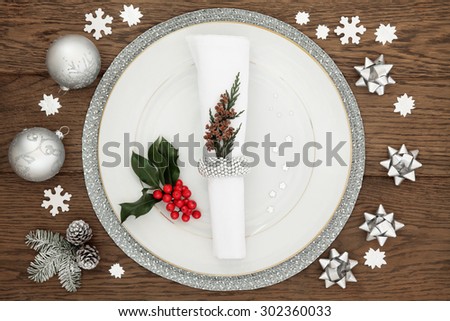 Christmas dinner time with plate, napkin, sparkling silver bauble decorations, holly and winter greenery over oak table background.