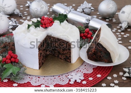 Christmas cake and slice with holly, bauble decorations and winter greenery over oak background.
