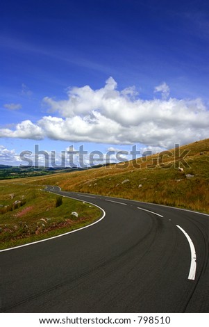 A rural mountain road with a left hand bend with puffy white clouds in a blue sky.