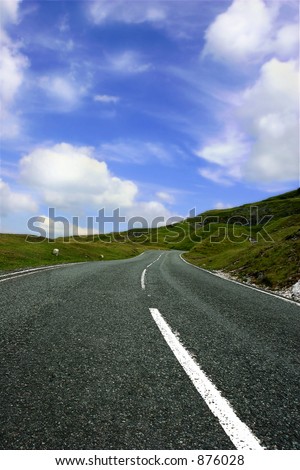 A steep, uphill  rural mountain road, with grass verges on either side and a blue sky with clouds.