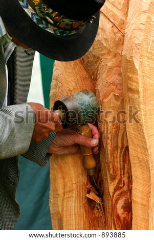 An elderly man in a suit with a hat on, using a chisel and mallet to carve a piece of wood.