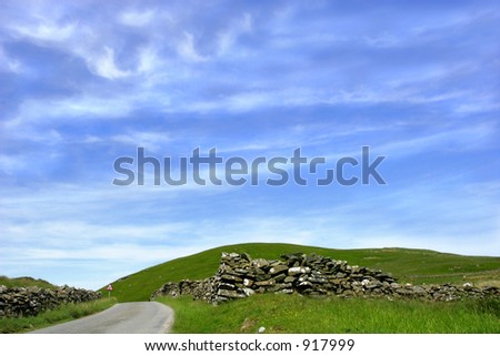 A mountain road with stone walls on either side and a blue sky and clouds.