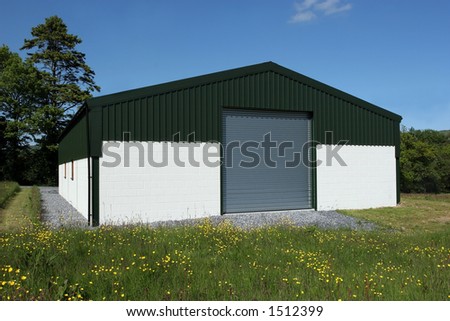 Newly  constructed barn of cream painted concrete block walls with a green metal sheet roof and roller shutter doors, standing in a field of buttercups, with a blue sky and trees to the rear.