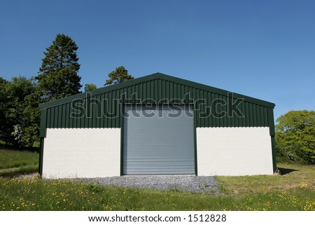 Newly  constructed barn of cream painted concrete block walls with a green metal sheet roof and roller shutter doors, standing in a field of buttercups, with a blue sky and trees to the rear.