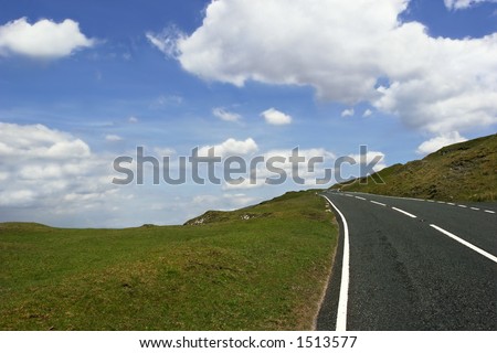 Uphill road in deserted countryside with grass verges either side on a blue sky with clouds. Set in the Brecon Beacons National Park, Wales, United Kingdom.