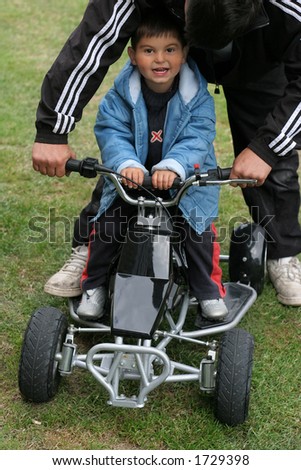 Little boy on a mini moto quad bike with an adult holding the handlebars and guiding the bike.