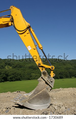 Steel bucket of an earth excavator standing idle on hardcore with trees and a blue sky to the rear.